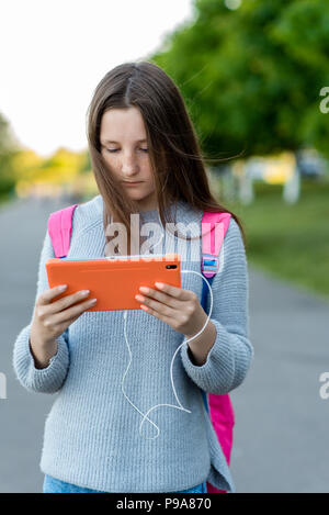 Girl schoolgirl teenager. Summer in nature. In her hands holds a tablet. Listen to music and watch the movie in the headphones. Concept of children on vacation after school. Behind the backpack. Stock Photo