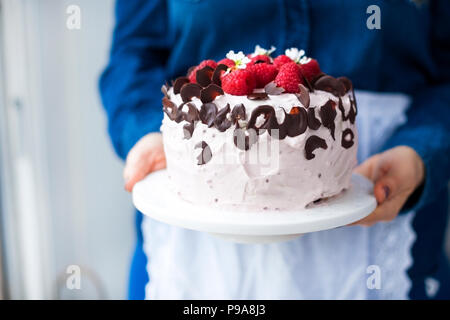 A woman is holding a beautiful cake with pink cream and fresh raspberry berries, decorated with chocolate. Calorie food. Free space for text or postcard