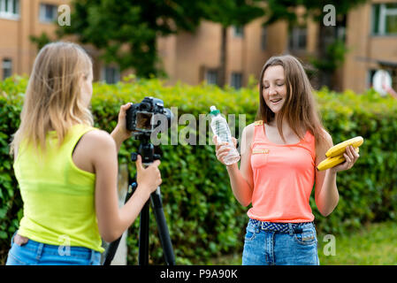 For girl schoolgirls, summer in nature. Records a video about healthy eating. In the hands holding a banana bottle with water. Use camera with tripod. Stock Photo