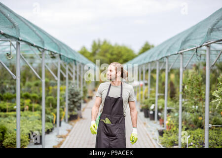 Handsome worker in uniform walking at the greenhouse with green plants for sale Stock Photo