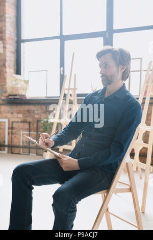Warm toned full length portrait of pensive mature man sitting alone in art studio and drawing sketches Stock Photo