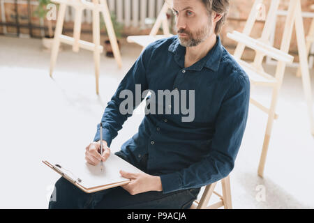 Warm toned  high angle portrait of pensive mature man sitting alone in loft space and drawing sketches Stock Photo
