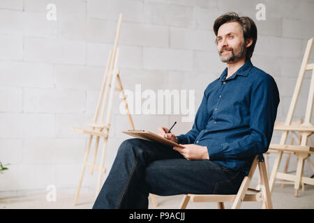 Warm toned portrait of smiling mature man sitting alone in loft space and drawing sketches, copy space Stock Photo