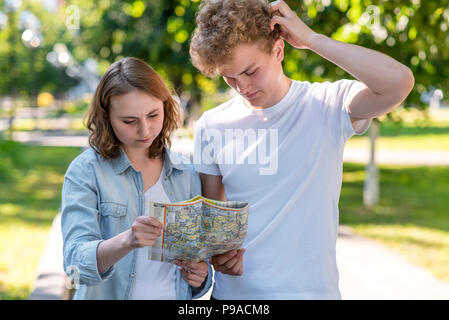 A guy with a girl in summer in the city on nature. In his hands holds a road map. Lost the direction. He does not know where to go. The concept of misunderstanding. He does not know where to go. Stock Photo