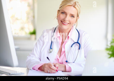 Close-up portrait of senior female doctor sitting at doctor's office at desk in front of laptop and doing some paperwork. Stock Photo