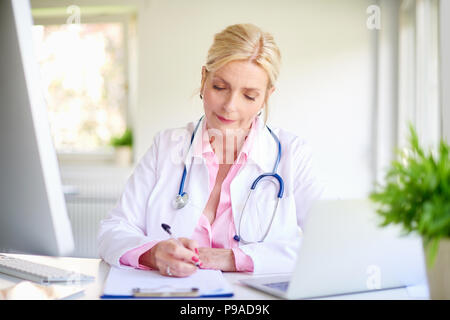 Close-up shot of smiling elderly female doctor using her laptop and writing something while sitting at desk at the doctor's office. Stock Photo