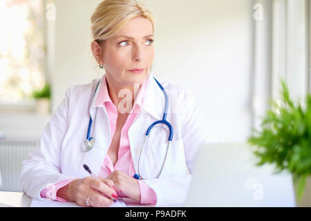 Close-up portrait of senior overwhelmed female doctor sitting at medical room and looking thoughtfully while writing something. Stock Photo