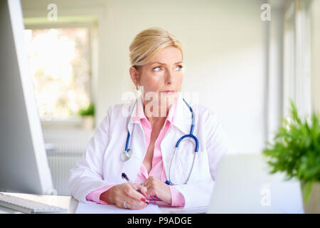 Close-up portrait of senior overwhelmed female doctor sitting at medical room and looking thoughtfully while writing something. Stock Photo