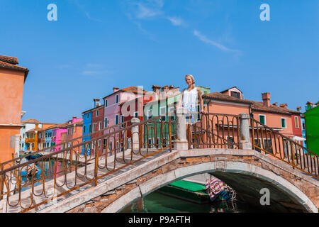 A female traveler stands on a bridge across a canal on the island of Burano with many colorful houses in the background Stock Photo