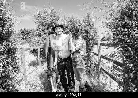 Cowboy and cowgirl, laughing. Male with open shirt and pecs as they are walking together with horse and saddle nearby. Stock Photo