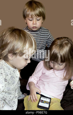 Little cute curious blonde triplets watch cartoons on smartphone indoors, half resaturated image isolated on dark beige Stock Photo