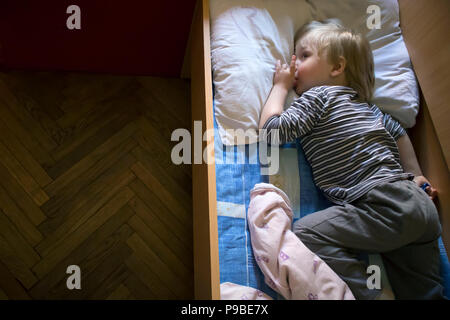 Little cute pensive blonde boy lying in bed and holds his finger in mouth Stock Photo