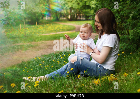 Young pregnant girl sits on lawn in city park. Mother holds her son on her knees. Little boy is happy. Blur effect Stock Photo