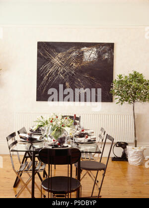 Black chairs and glass table set for Christmas dinner in modern white dining room with abstract painting above bay tree in white pot Stock Photo