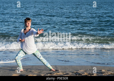 Young woman with brown hair wearing white blouse standing on a beach by the ocean, doing Tai Chi. Stock Photo