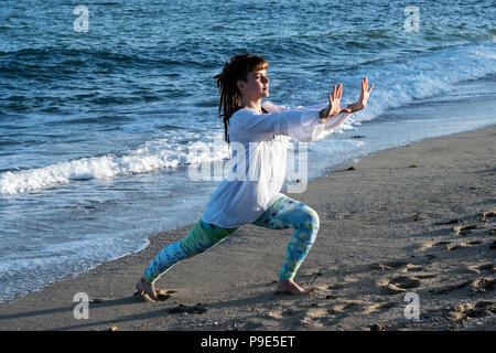 Young woman with brown hair wearing white blouse standing on a beach by the ocean, doing Tai Chi. Stock Photo