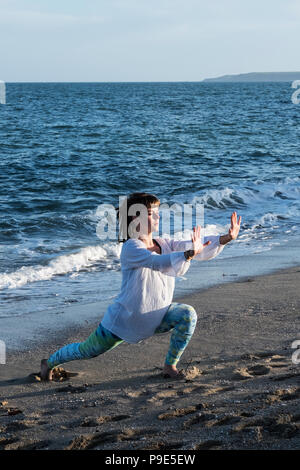 Young woman with brown hair wearing white blouse standing on a beach by the ocean, doing Tai Chi. Stock Photo