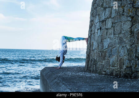 Young woman doing handstand by the ocean, with her legs against a wall. Stock Photo