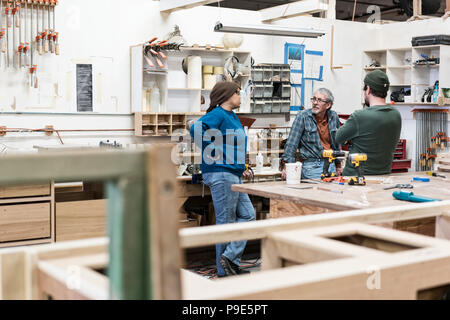 A group of mixed race carpenters discussing a project at a work station in a large woodworking shop. Stock Photo