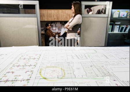 Closeup of plans on a table with two people working in the background. Stock Photo