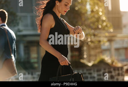 Woman looking at her wrist watch while commuting to office in the morning carrying her bag. Office going people walking on a street in the morning. Stock Photo