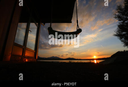 12 July 2018, Germany, Fuessen: The sun sets behind a swing at a boathouse at Hopfensee lake. Photo: Karl-Josef Hildenbrand/dpa Stock Photo