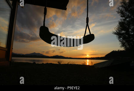 12 July 2018, Germany, Fuessen: The sun sets behind a swing at a boathouse at Hopfensee lake. Photo: Karl-Josef Hildenbrand/dpa Stock Photo