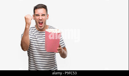 Handsome young man eating popcorn annoyed and frustrated shouting with anger, crazy and yelling with raised hand, anger concept Stock Photo