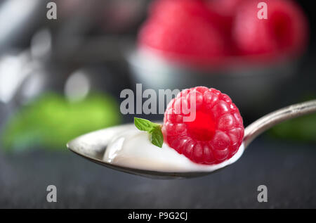 Close-up on a spoon of yougurt with juisy raspberry on top with berries and meant leaves out of focus Stock Photo