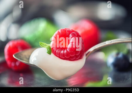 Close-up on a spoon of yougurt with juisy raspberry on top with berries and meant leaves out of focus Stock Photo