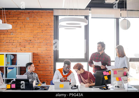 thoughtful Indian guy holding paper and looking away in the office with colleagues Stock Photo