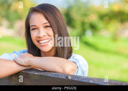 Beautiful happy Asian Eurasian young woman or girl wearing denim shirt, smiling with perfect teeth and leaning on fence in sunshine Stock Photo