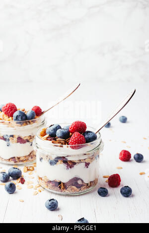 Two jars with tasty parfaits made of granola, berries and greek yogurt on white wooden table. Shot at angle. Stock Photo