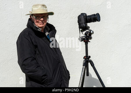 Senior male wearing spectacles,Tilley Hat and outdoor coat standing by his tripod and camera against a white wall Stock Photo