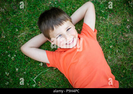 Cute happy 6 years old boy lying on green grass and smiling. Stock Photo