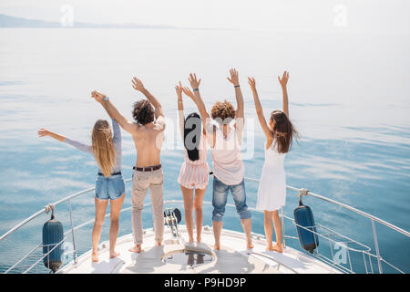 Rear view of cheerful caucasian teenagers on a sailing boat, lifting their hands up, looking at blue sea surfice, full of happiness,celebrating the st Stock Photo