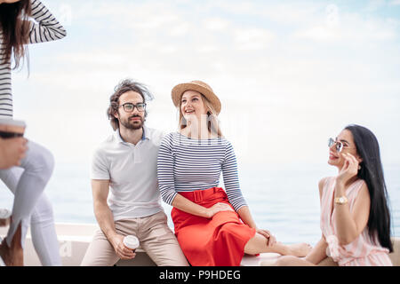 European attractive brown-haired man in spectaces and blonde woman in straw hat and stripped blouse, embracing, chilling with their friends at sailing Stock Photo
