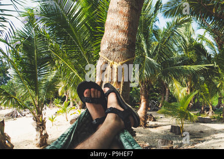 Men's legs in shorts lying in a hammock on the sand beautiful tropical beach in the shade of palm trees. Maldives. Stock Photo