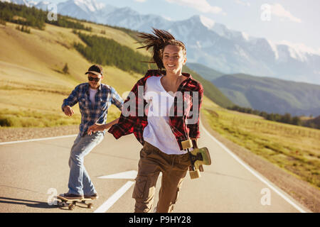 Happy friends are having fun with skates and longboards at straight road against mountains Stock Photo