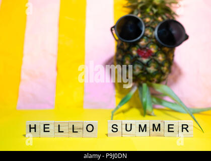 Wooden cubes with written words Hello summer and red lip pineapple wears sunglasses, having sun bath on bright yellow and pink pastel stripe backgroun Stock Photo
