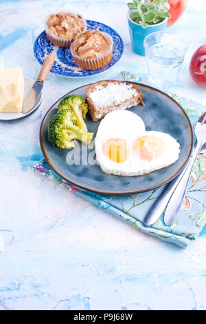 Fried eggs in the form of heart. On a dark plate with broccoli and toast. Blue cup with tea, apples, butter for breakfast and cupcakes. On a light colored background. Free space for text or advertisin. Stock Photo