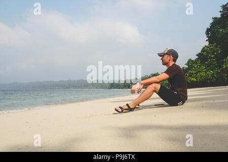 a young guy sits on a white sandy beach and looks at the sea on an exotic island. Man enjoying on empty tropical exotic beach. tourist visitor travell Stock Photo