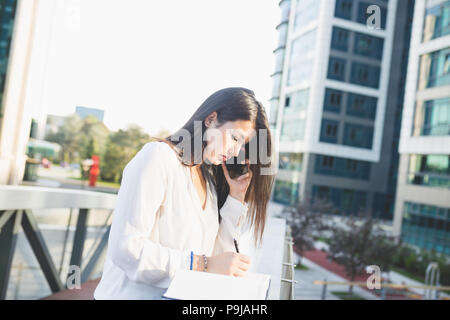 Business woman portrait outdoors talking at the phone and writing notes, with modern building in background. Stock Photo