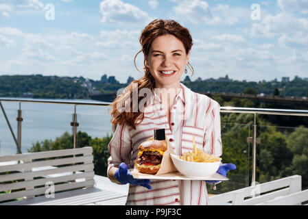 Smiling woman with ponytail eating potatoes and meat burger Stock Photo