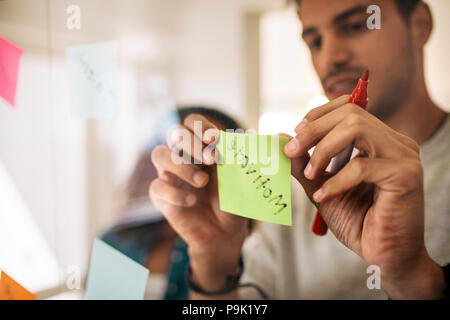 Man writing on sticky notes with marker pen and pasting on glass board in office. Entrepreneurs discussing business ideas and plans with sticky notes  Stock Photo