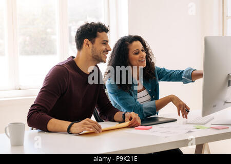 Businesswoman pointing towards the computer with a digital writing pad on the table. Woman entrepreneur discussing work with colleague sitting at her  Stock Photo