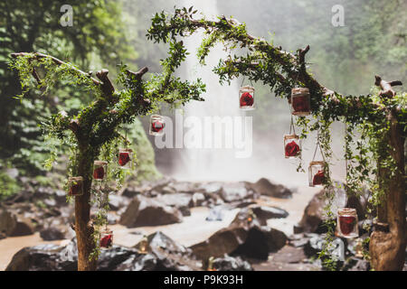 Tropical wedding ceremony with waterfall view in jungle canyon. Decorated with green ivy, old branches and hanging lamps Stock Photo