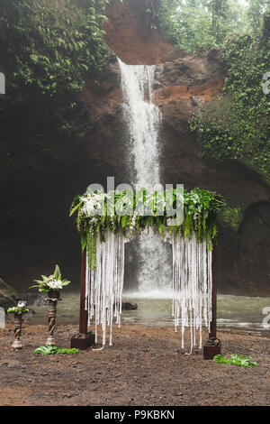 Tropical wedding ceremony with waterfall view. White arch decorated with green jungle leaves monstera and fern Stock Photo