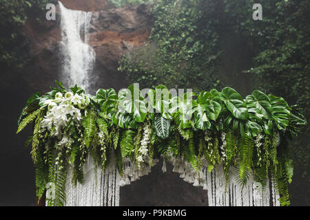 Tropical wedding ceremony with waterfall view. White arch decorated with green jungle leaves monstera and fern Stock Photo