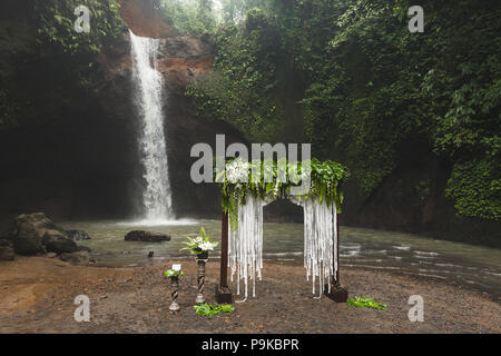 Tropical wedding ceremony with waterfall view. White arch decorated with green jungle leaves monstera and fern Stock Photo